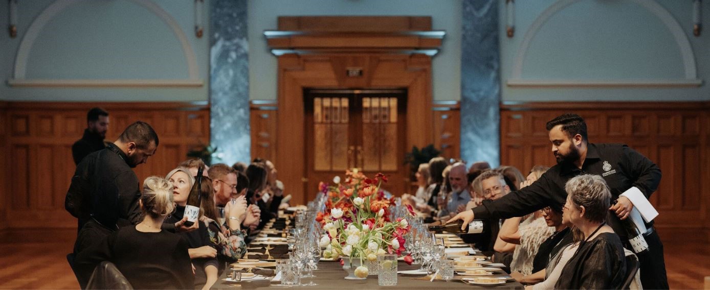 photo from end of long table during formal dinner service with decorative floral arrangements at table centre.  Guests and service staff can be seen around table.