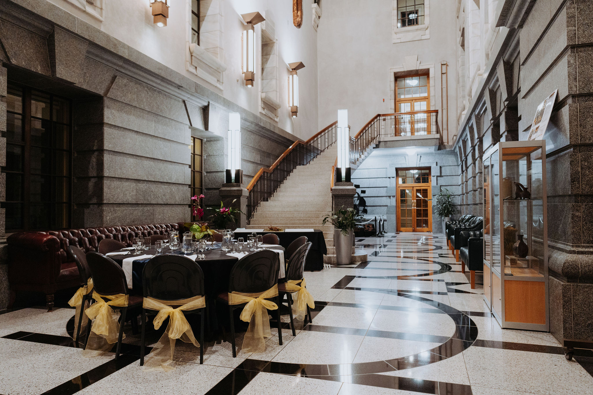 photo of table setup for dinner in Galleria space. Gold ribbons decorate the chairs and white, black and grey marble can be seen on the floor and walls