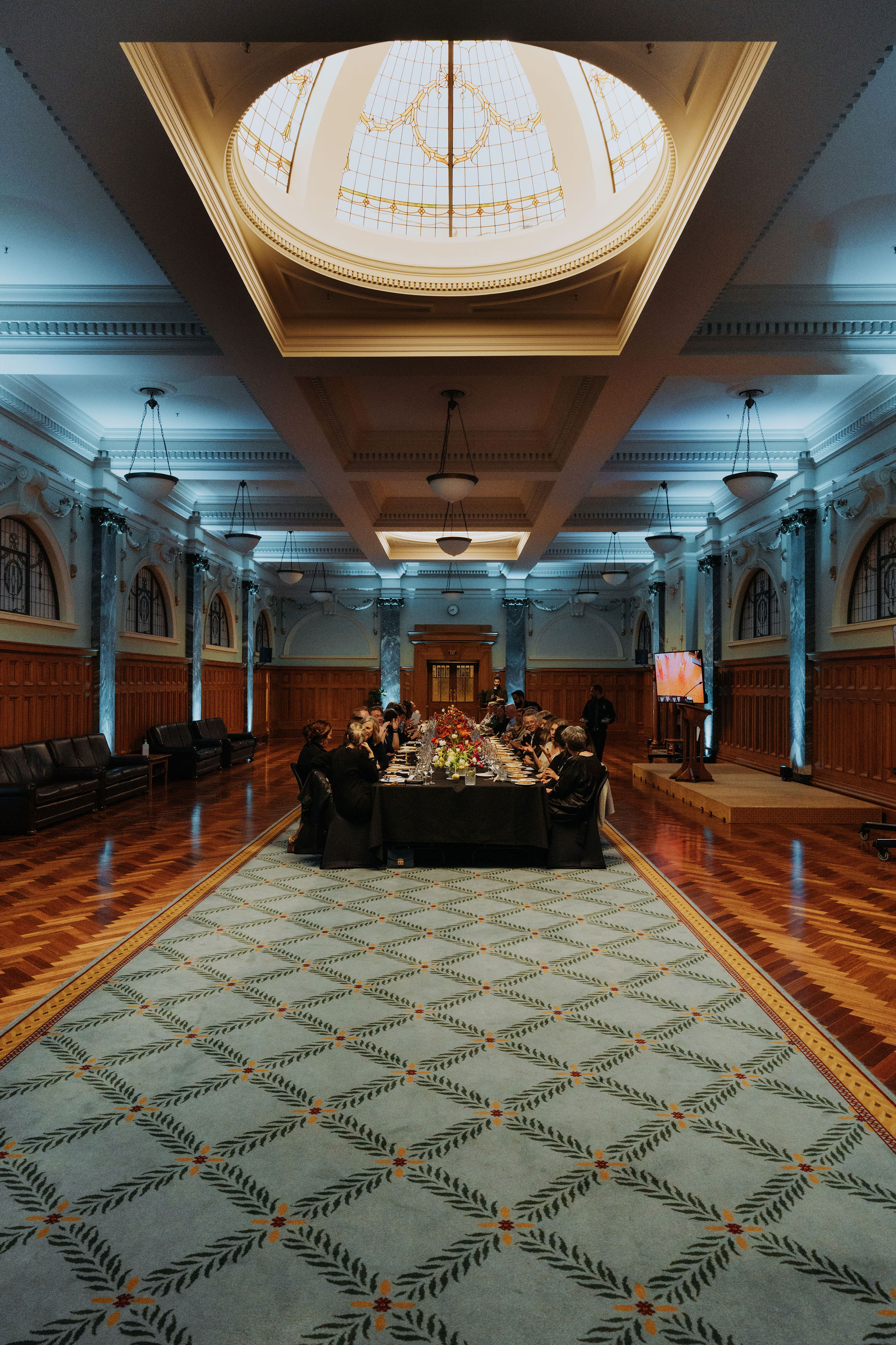 wide photo from end of long table setup for dinner in Grand Hall with guests seated. A stage with TV screens can be seen to one side.