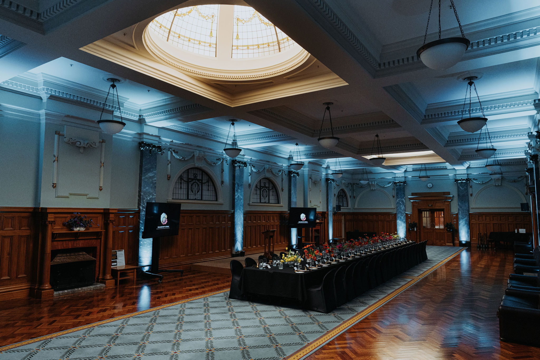 wide photo of long table setup for dinner in Grand Hall with stage in background with TV screens on either side