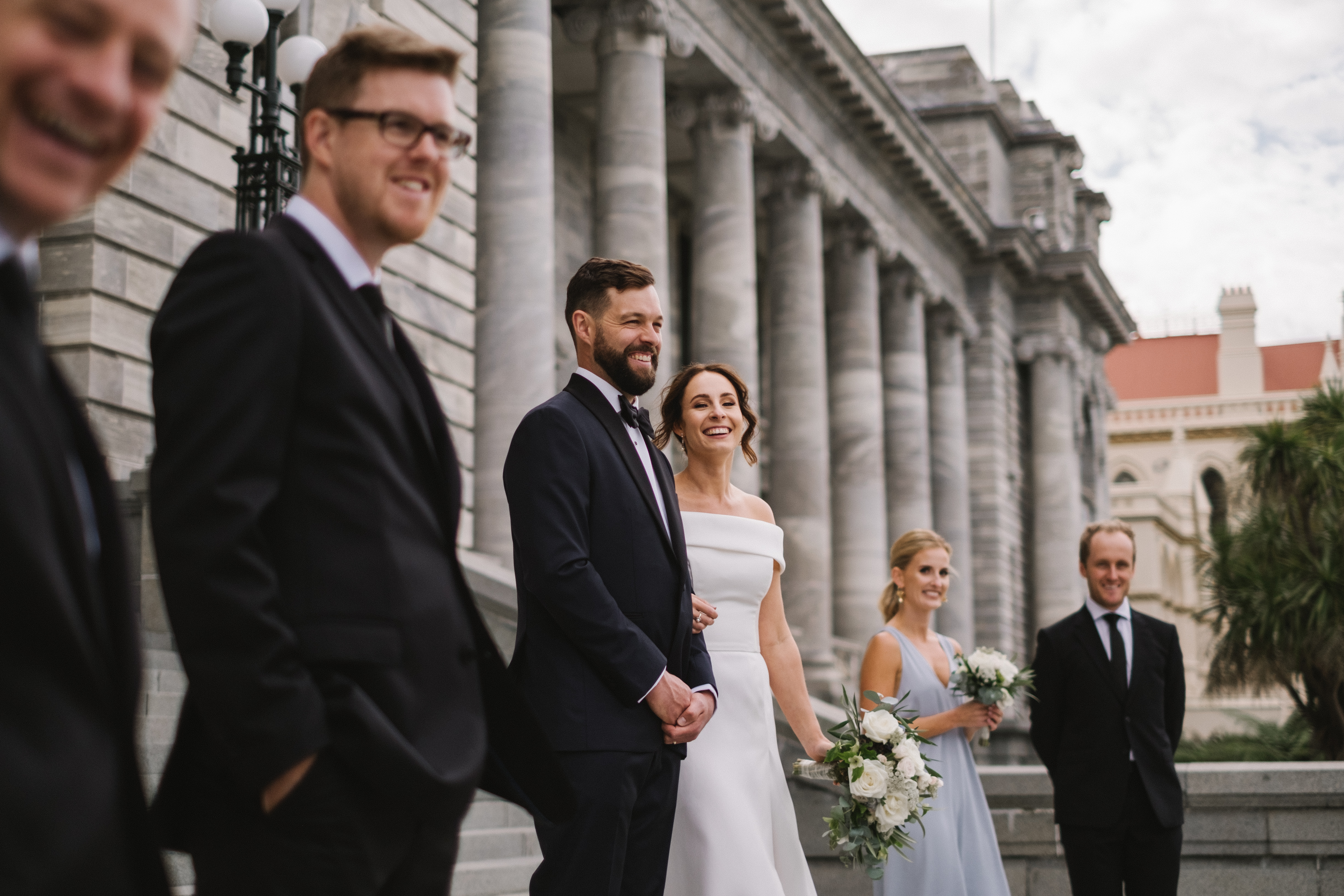 photo of couple dressed in wedding attire with some wedding guests either side, standing outside parliament buildings..