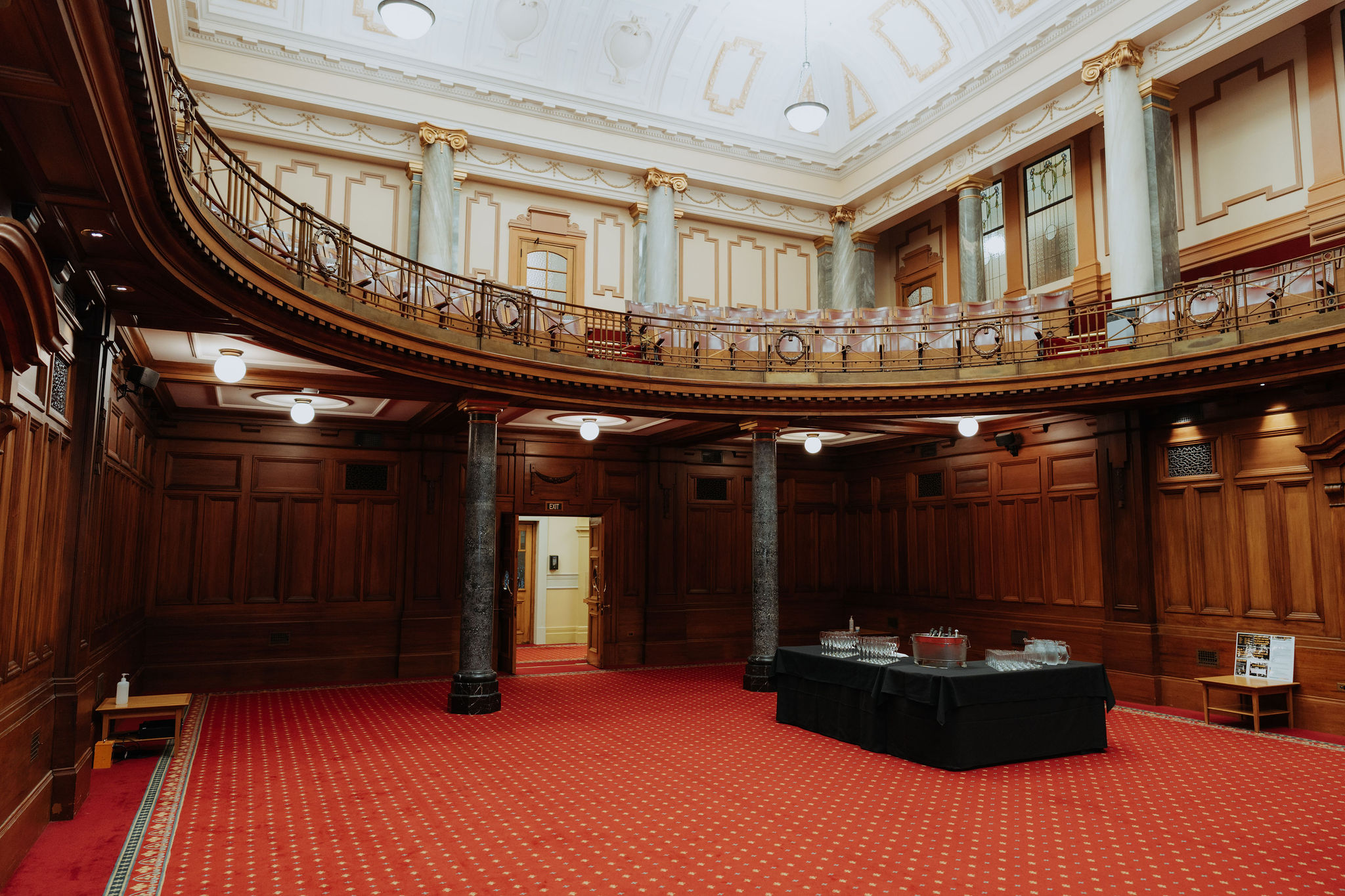 wide photo of the Leglislative Council Chamber showing balcony level and red carpet. A table for drinks can be seen setup to one side.