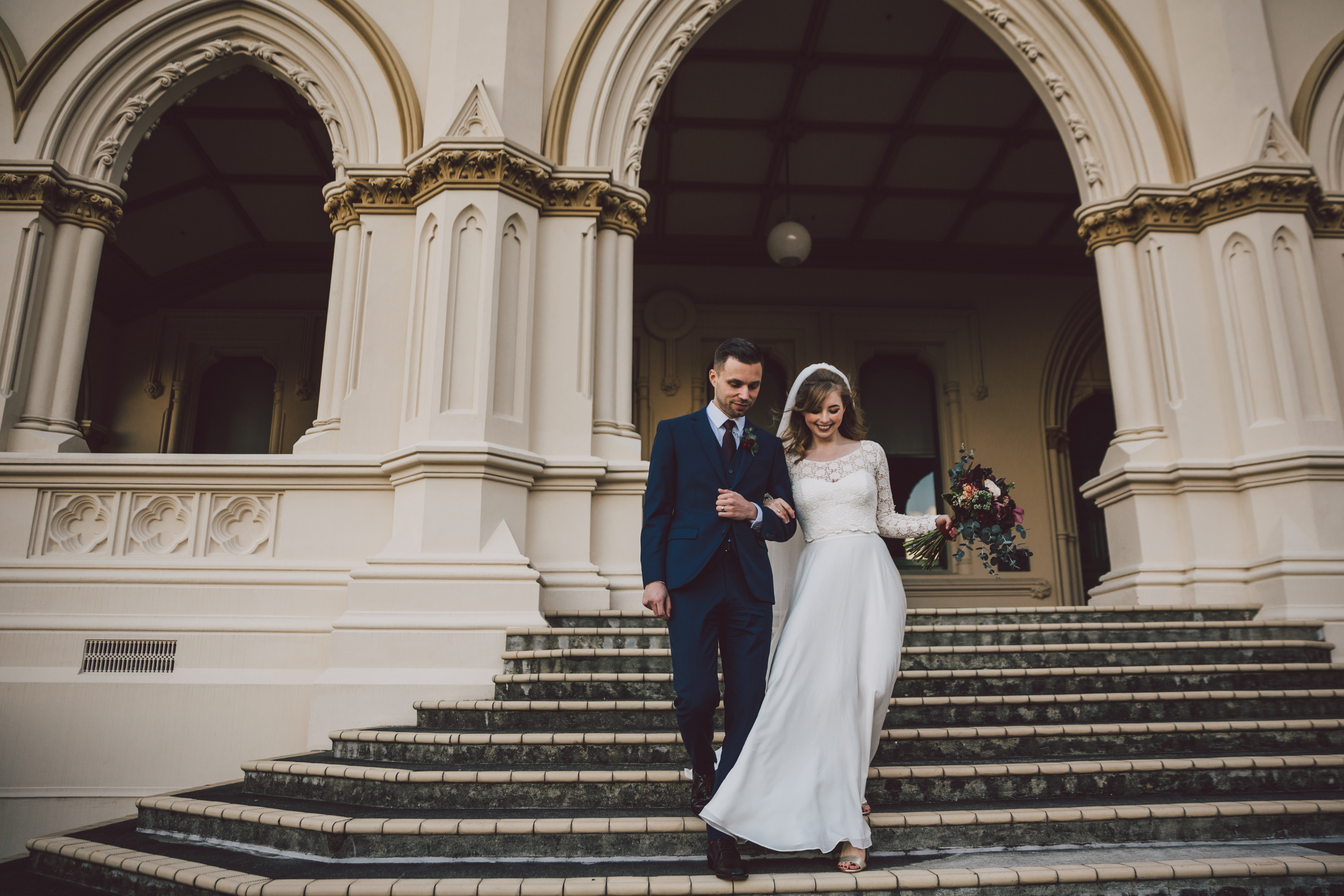 photo of a couple dressed in wedding attire, walking down steps of ornate building. .