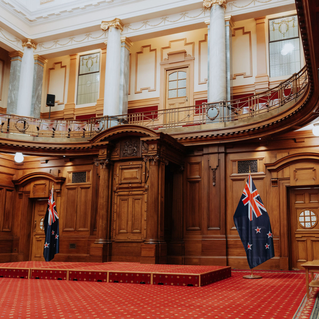 photo showing ornate architecture with carved wood panels and marble pillars. A small stage can be seen with a New Zealand flag on either side.